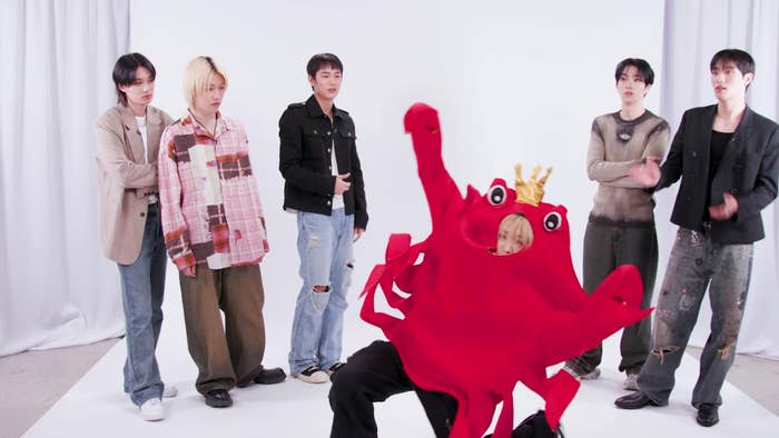 Five people chat while one person in a red crab costume with a crown waves. Everyone is facing the person in the costume