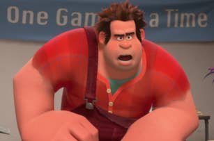 Ralph from Wreck-It Ralph stands with an angry expression, wearing a red plaid shirt and brown overalls, with a banner reading "One Game at a Time" behind him