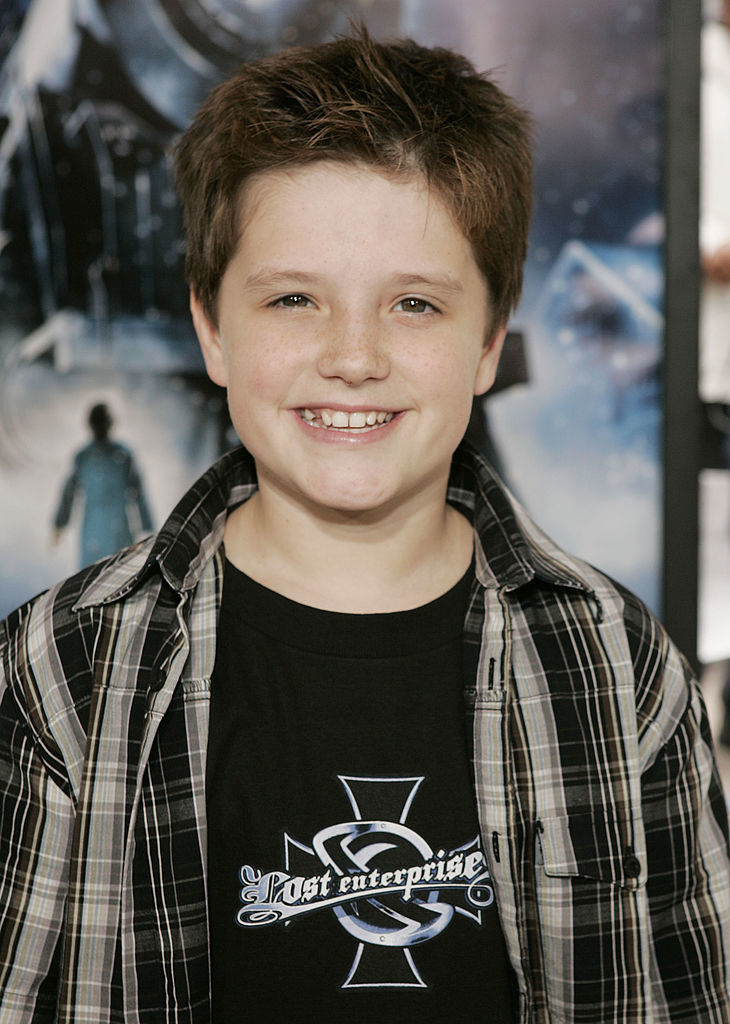 Young boy smiling, wearing a plaid shirt over a t-shirt with the logo &quot;Lost Enterprises,&quot; at a media event. Name not provided or recognizable