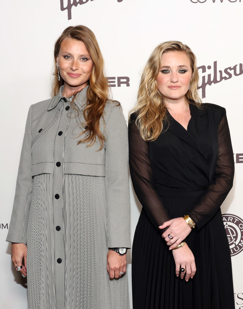 Aly Michalka and AJ Michalka pose on the red carpet. Aly is wearing a checkered coat, and AJ is in a black dress with sheer sleeves