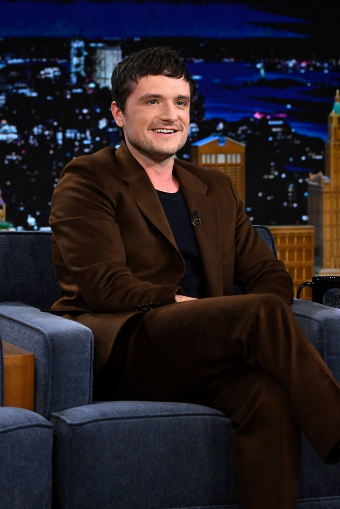 Josh Hutcherson, seated and smiling, in a brown suit and black shirt on a talk show set with a cityscape backdrop