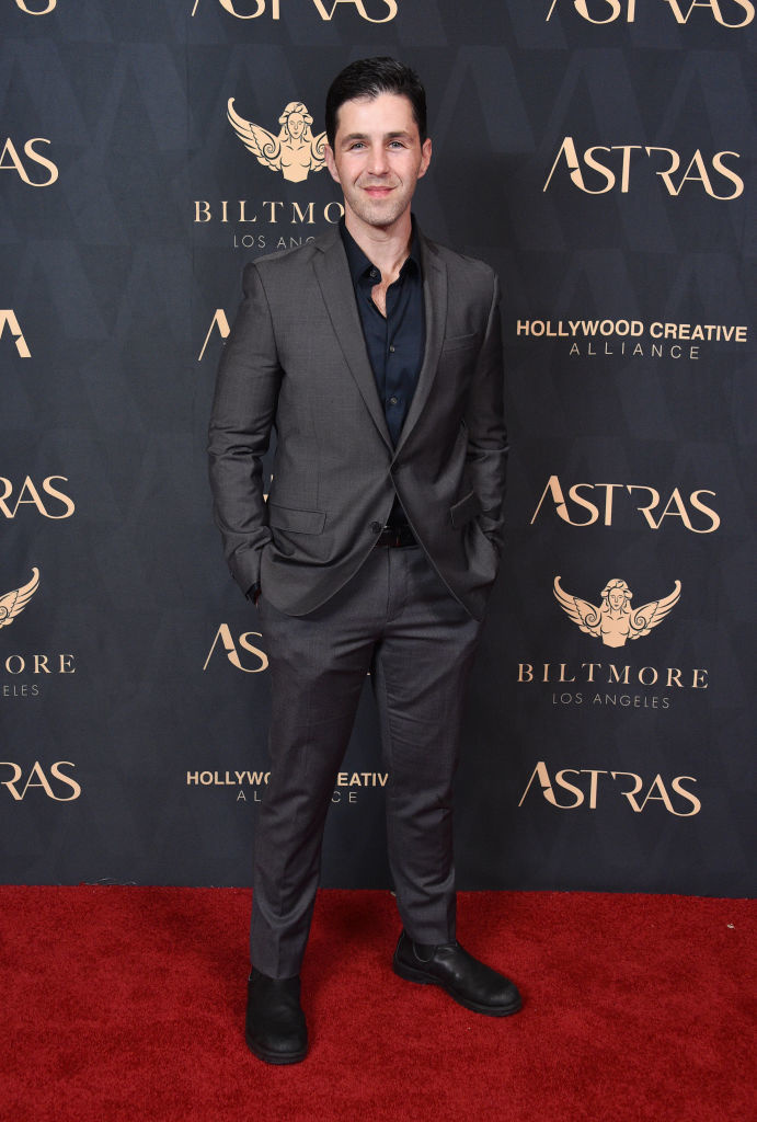 Josh Peck on the red carpet at the Hollywood Creative Alliance event, wearing a dark suit and black shirt. Background features Biltmore Los Angeles and ASTRAS logos