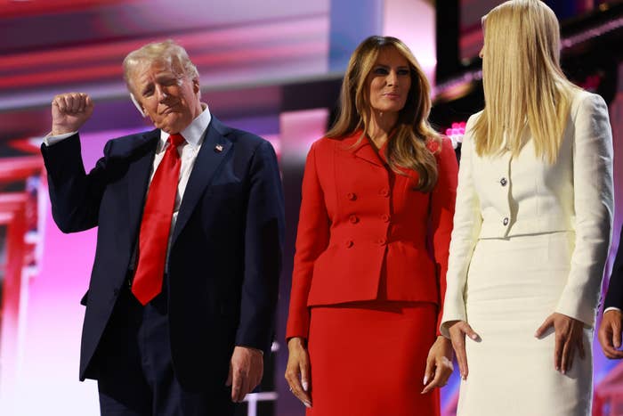 Donald Trump, Melania Trump, and Ivanka Trump stand together. onstage. Donald raises his fist, Melania wears a formal outfit, and Ivanka wears a formal dress