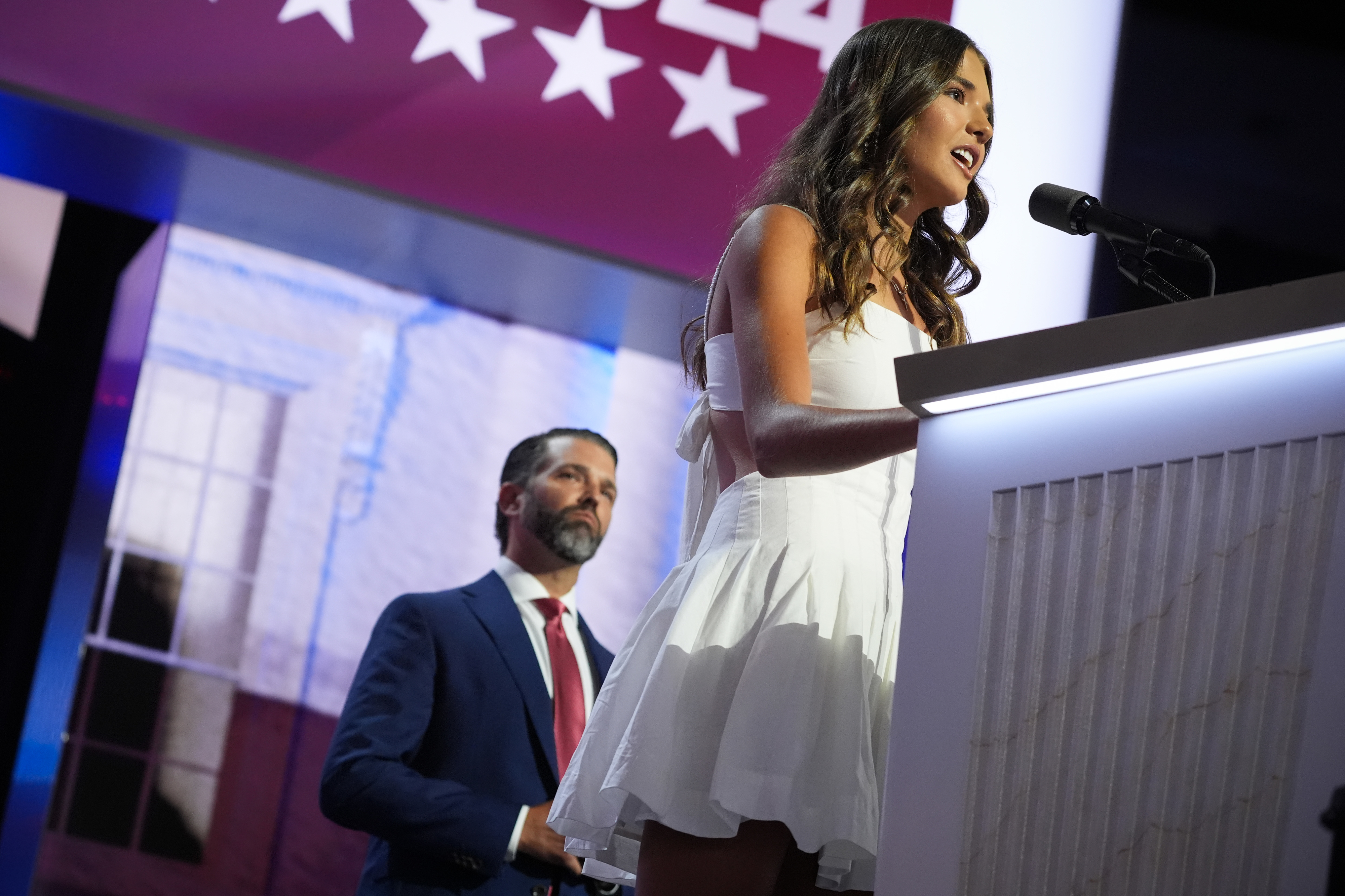 Kai Madison Trump speaks at a podium in a sleeveless dress, while Donald Trump Jr. stands behind her in a blue suit and red tie