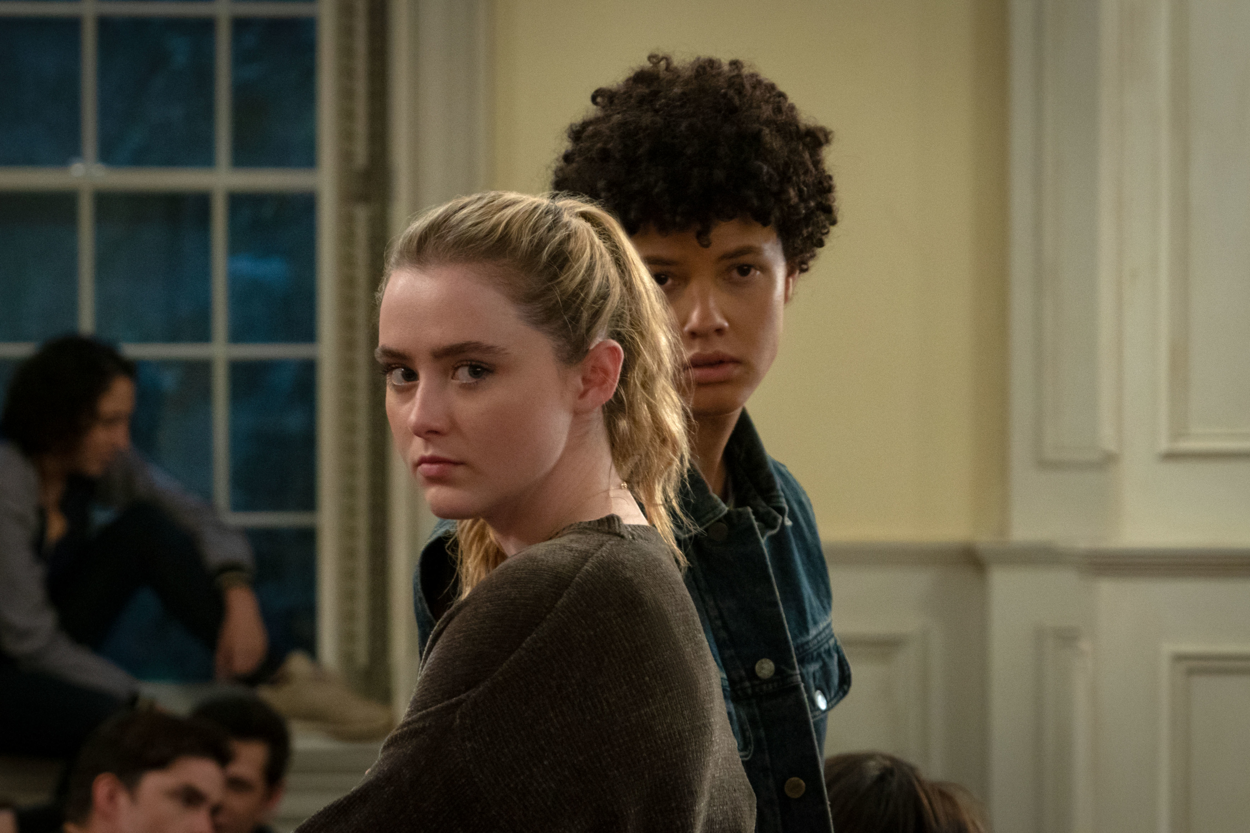 Kathryn Newton and a person with curly hair stand inside a room with serious expressions, several other people are seated in the background