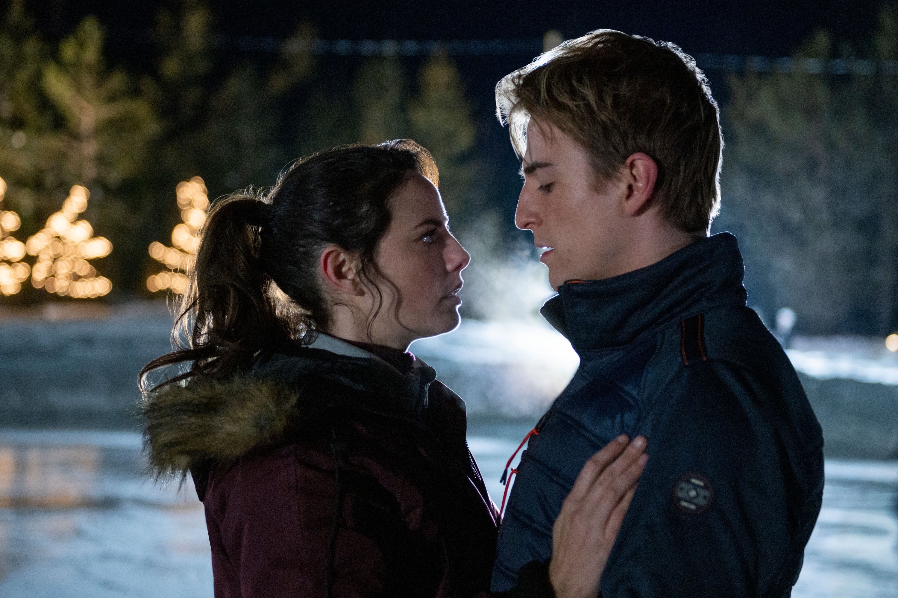 Kaya Scodelario and Evan Roderick standing outdoors in an ice rink at night, facing each other closely, with trees illuminated in the background