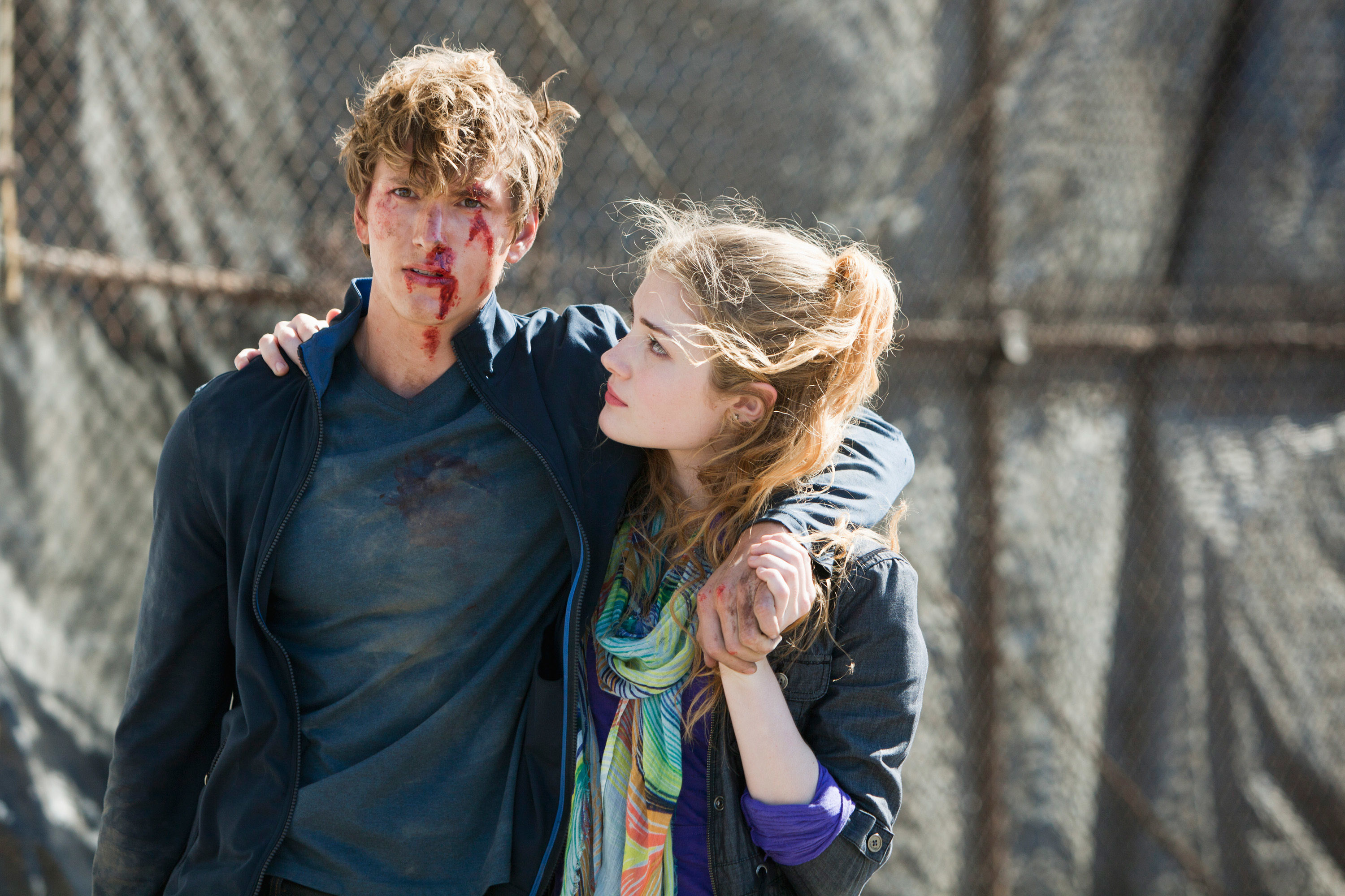 A young man with a bloodied face and disheveled hair, is supported by Skyler Samuels who holds his hand and looks at him empathetically