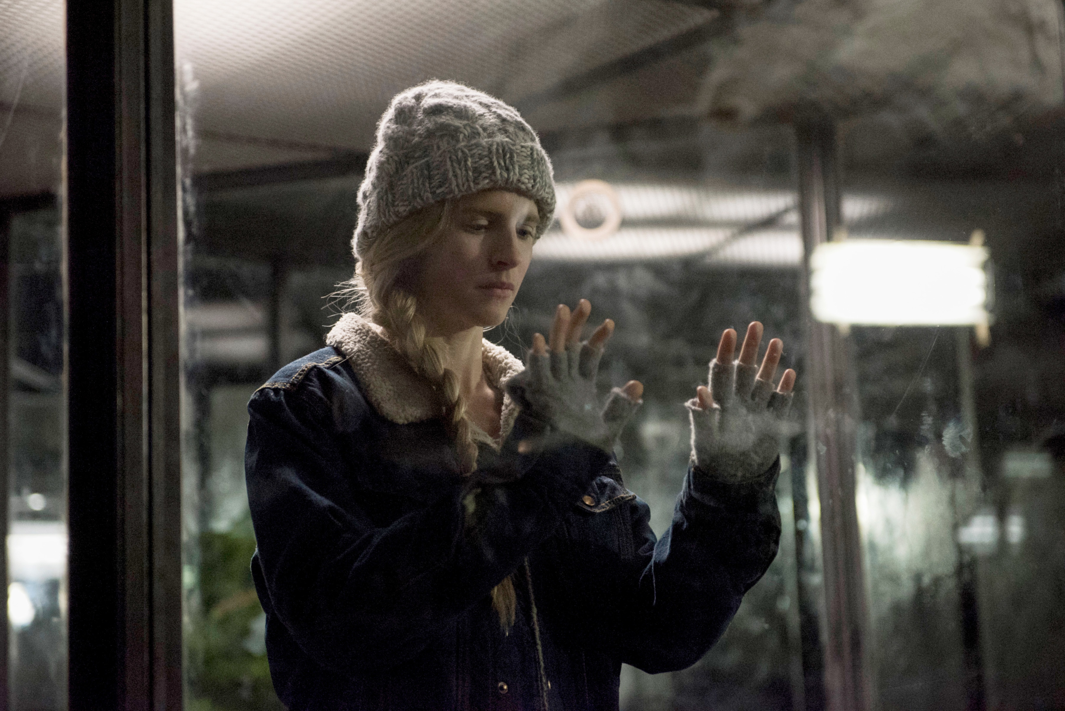 Brit Marling, wearing a knit hat and a jacket, touches a foggy glass window with both hands in a contemplative pose