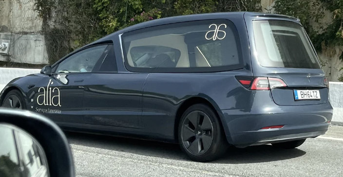 Blue hearse with &quot;alia&quot; business logos on the side and rear window, seen driving down a road