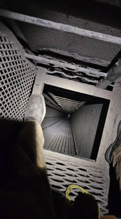 Two people at the top of a dark, narrow shaft with a metal grid floor, one person&#x27;s shoes and legs partially visible