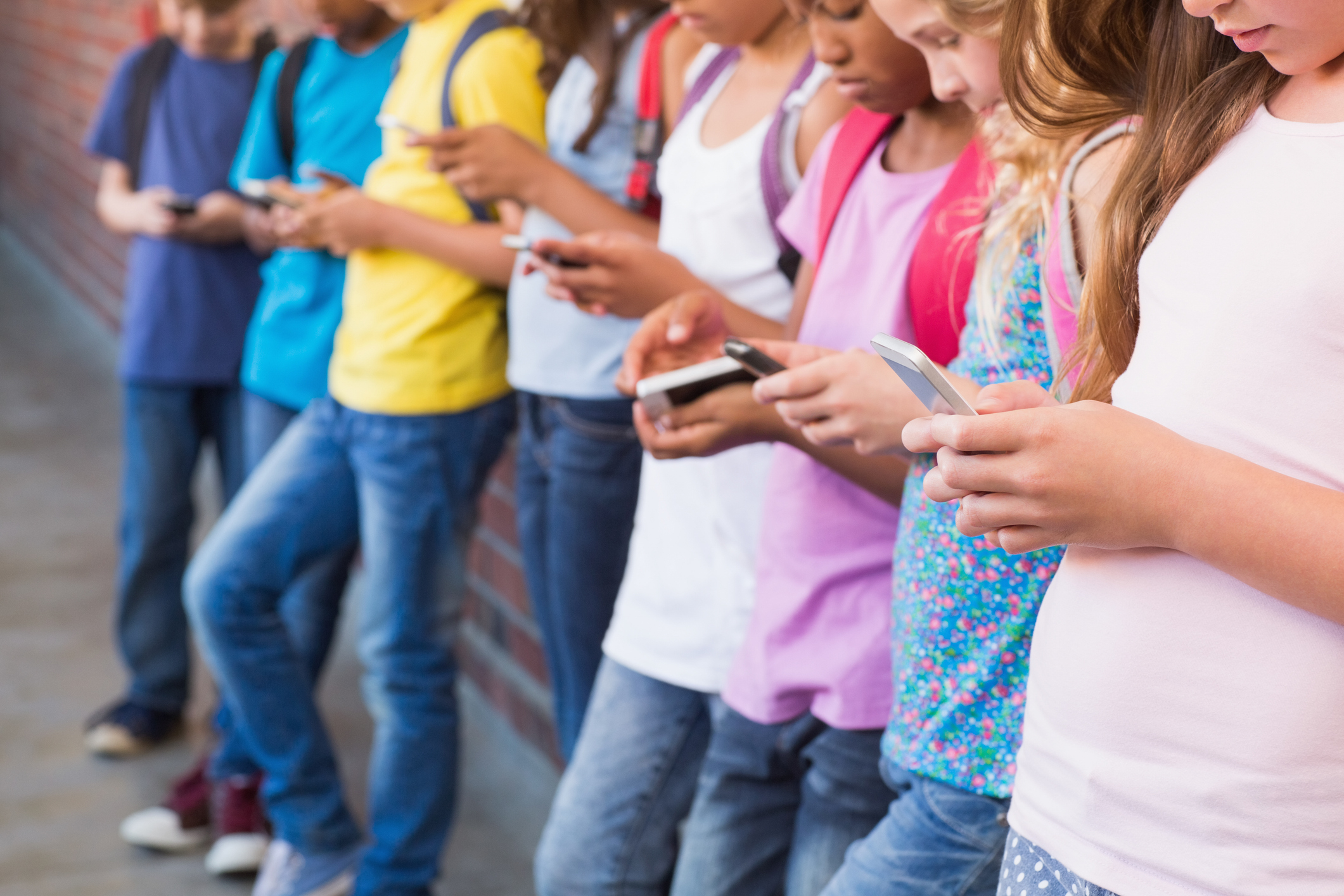 A group of children stand in a row against a brick wall, each focused on their smartphones. They wear casual clothing, including T-shirts and jeans