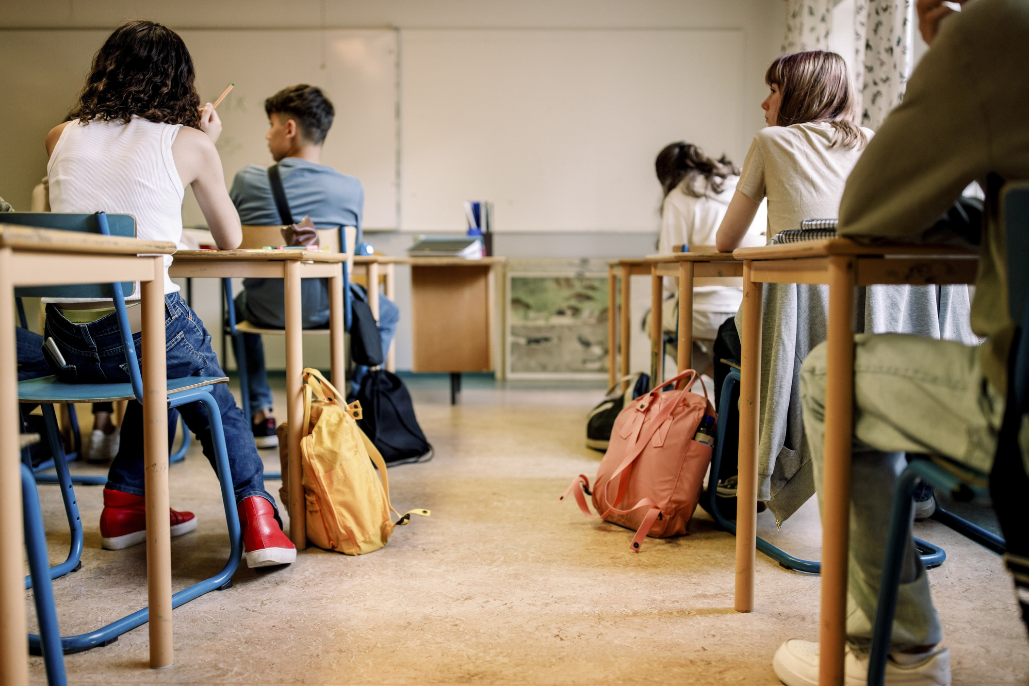 Students sit at desks in a classroom, facing the front with their backpacks on the floor beside them