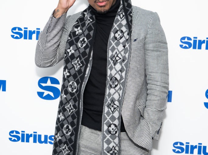 Nick Cannon poses in a checkered suit with a patterned scarf at a SiriusXM event backdrop, adjusting his sunglasses