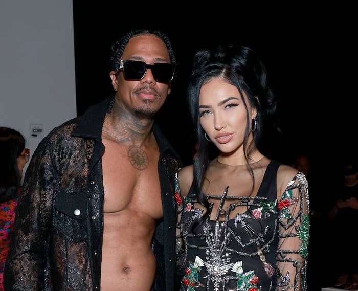 Nick Cannon and Bre Tiesi at a fashion event. Nick wears a sheer black shirt and sunglasses, while Bre wears an intricate sheer black dress with floral details