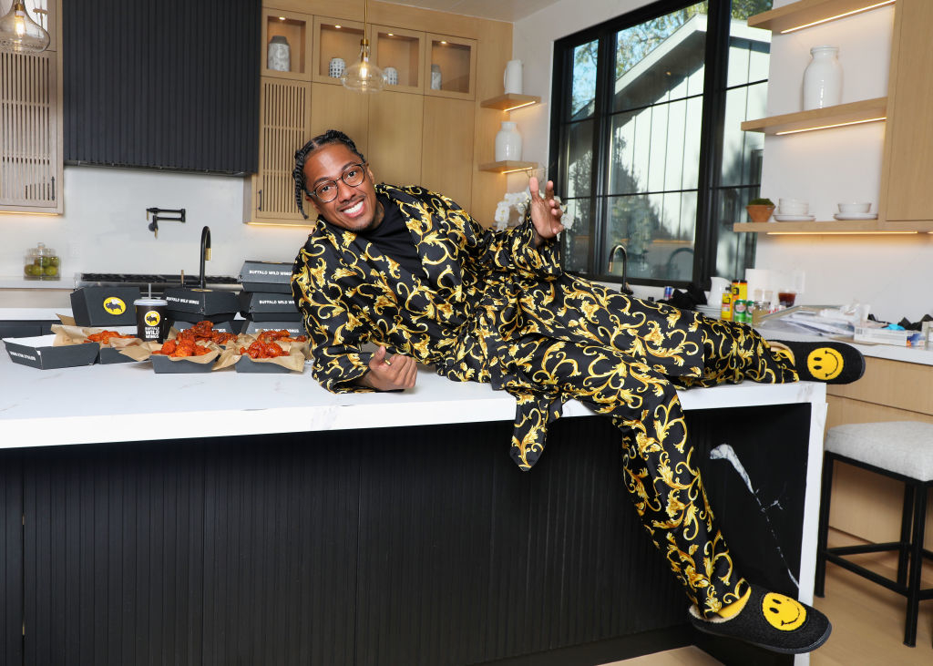 Nick Cannon is lounging on a kitchen counter, smiling and wearing a black and gold patterned suit with smiley face slippers. Boxes of wings are on the counter