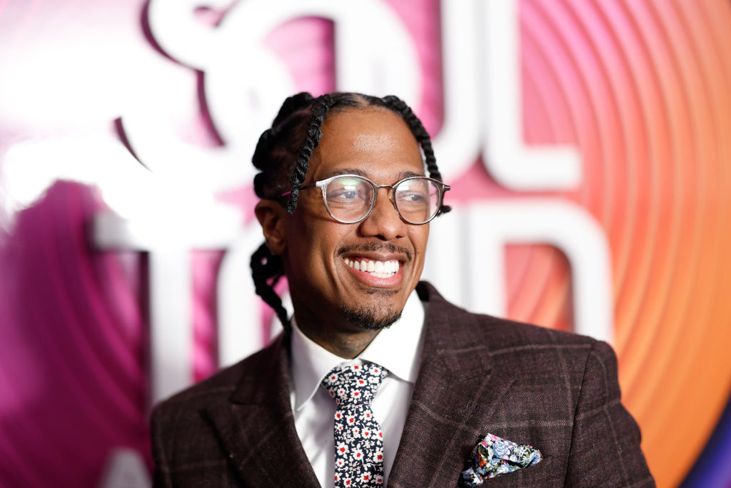 Nick Cannon smiling, wearing glasses, a dark plaid suit, and a floral tie, standing in front of a &quot;Soul Train Awards&quot; backdrop