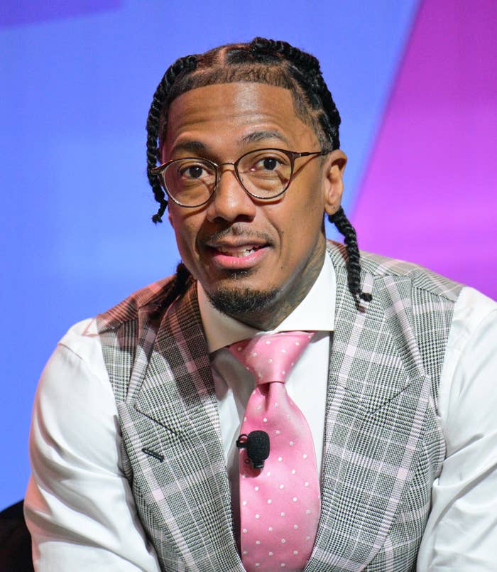 Nick Cannon sits, wearing glasses, a plaid suit vest, and a pink tie with polka dots. He has a serious expression and braided hair