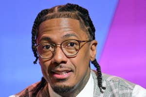 Nick Cannon sits, wearing glasses, a plaid suit vest, and a pink tie with polka dots. He has a serious expression and braided hair