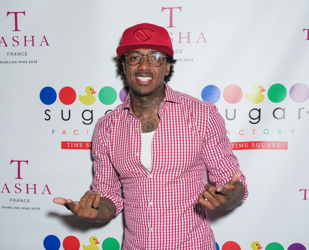 Nick Cannon wearing a red cap and a red checkered shirt, smiling and posing at a Sugar Factory event