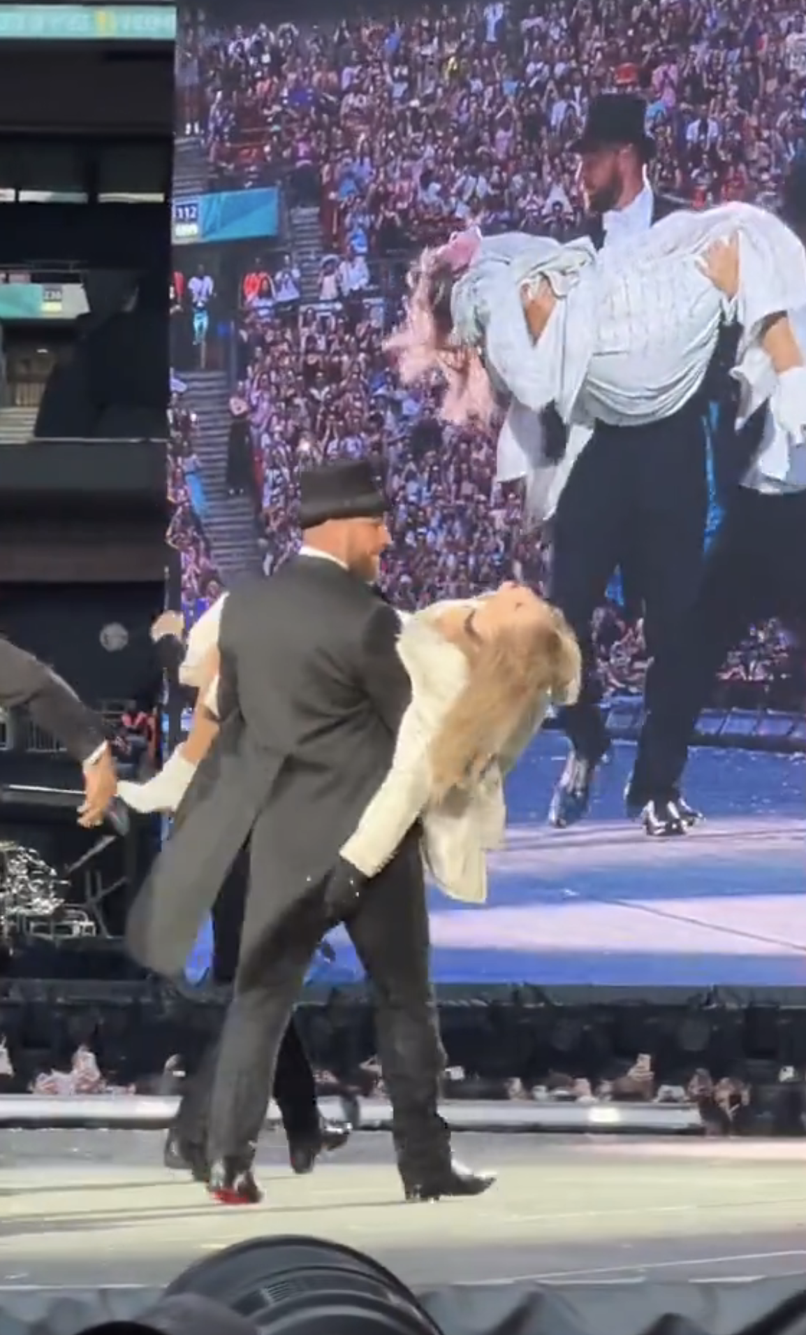 Travis Kelce carries Taylor Swift, who is pretending to be unconscious, during a concert, with a large screen showing the same scene in the background