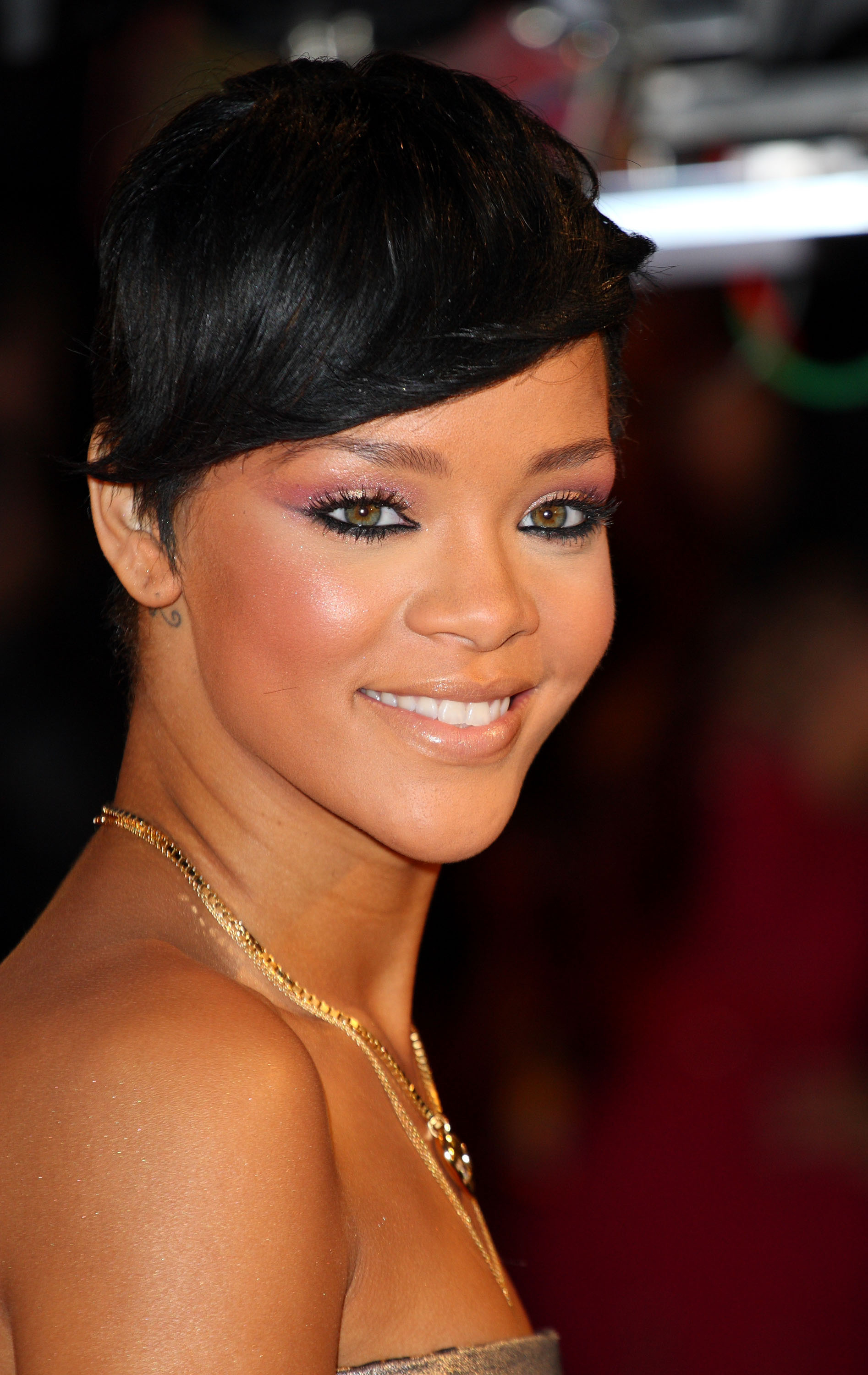 Rihanna is smiling on a red carpet with pale, concealer makeup coated lips, wearing a strapless dress and a gold necklace, with short, styled hair