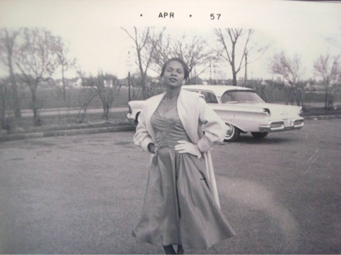 A woman in a vintage dress and coat poses in a parking lot with a classic car behind her. Photo dated April 1957