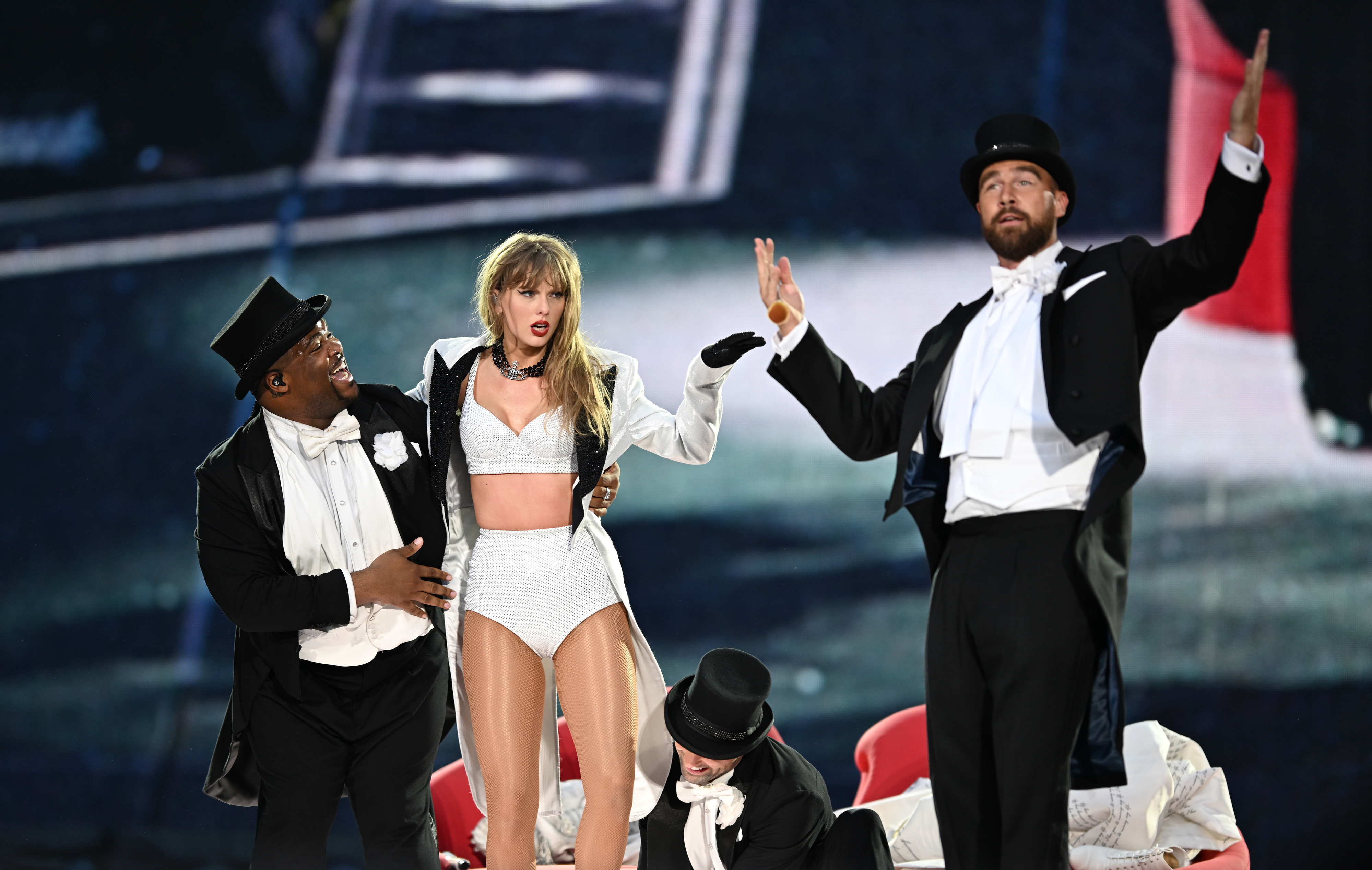 Taylor Swift, in a performance outfit, stands between two male dancers, including Travis, in tuxedos. Another dancer in a tuxedo kneels in front. They are on stage