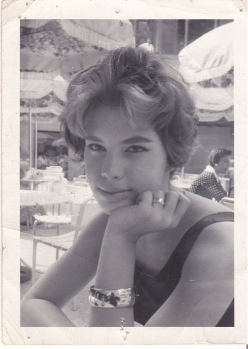 A woman with short hair, wearing a sleeveless top and bracelet, sits outdoors with her chin resting on her hand. Sun umbrellas and tables are in the background