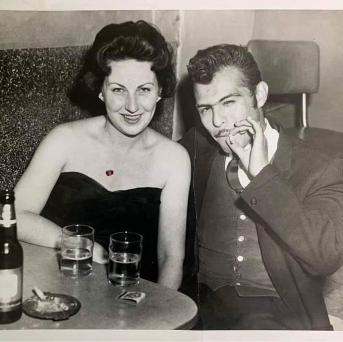 A man and woman sit together at a table with drinks, both smiling at the camera. The woman wears a strapless dress, and the man is in a suit, holding a cigarette