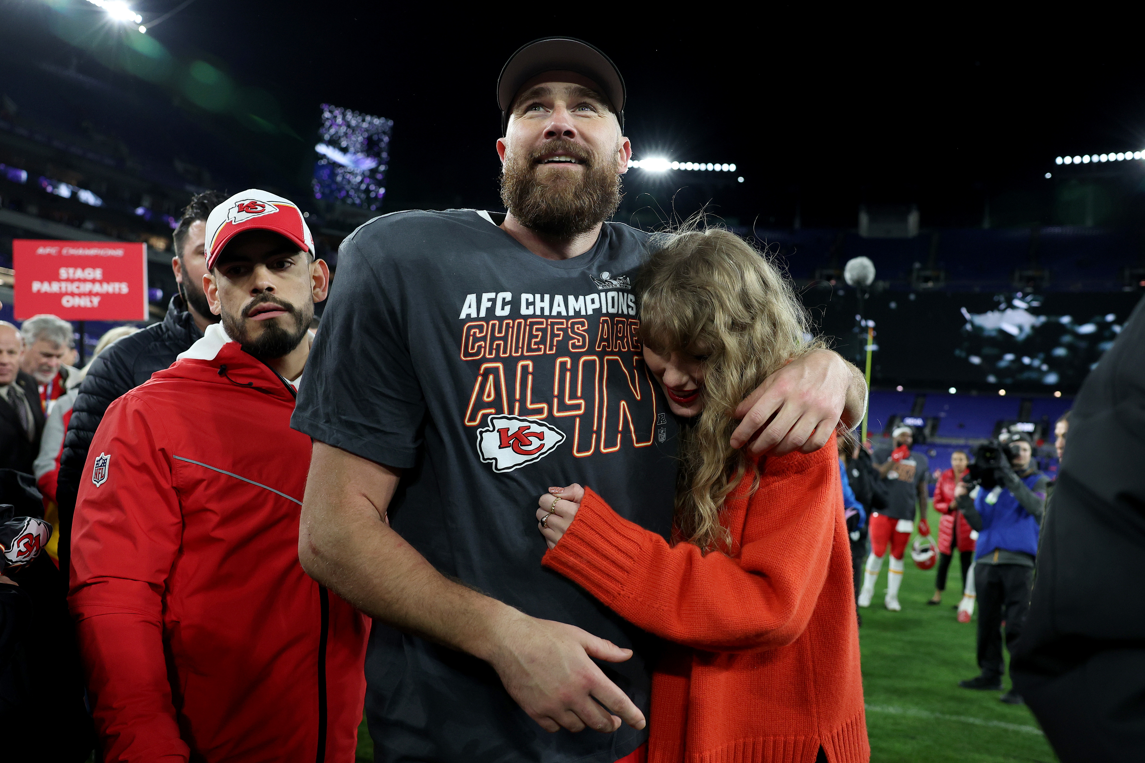 Travis Kelce, in casual sports clothing, embraces Taylor Swift, wearing a cardigan, on a football field. Another man in red sports gear stands near them