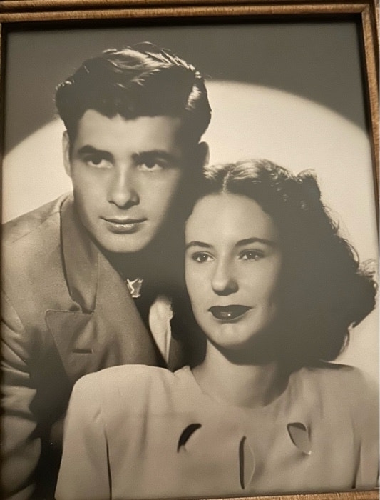 A black and white photograph of a man and a woman from the mid-20th century. The man is wearing a suit, and the woman is in a blouse with decorative cutouts at the neckline