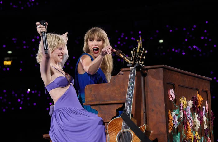 Hayley Williams and Taylor Swift perform on stage with a decorated piano and guitar. Both are smiling and wearing elegant dresses
