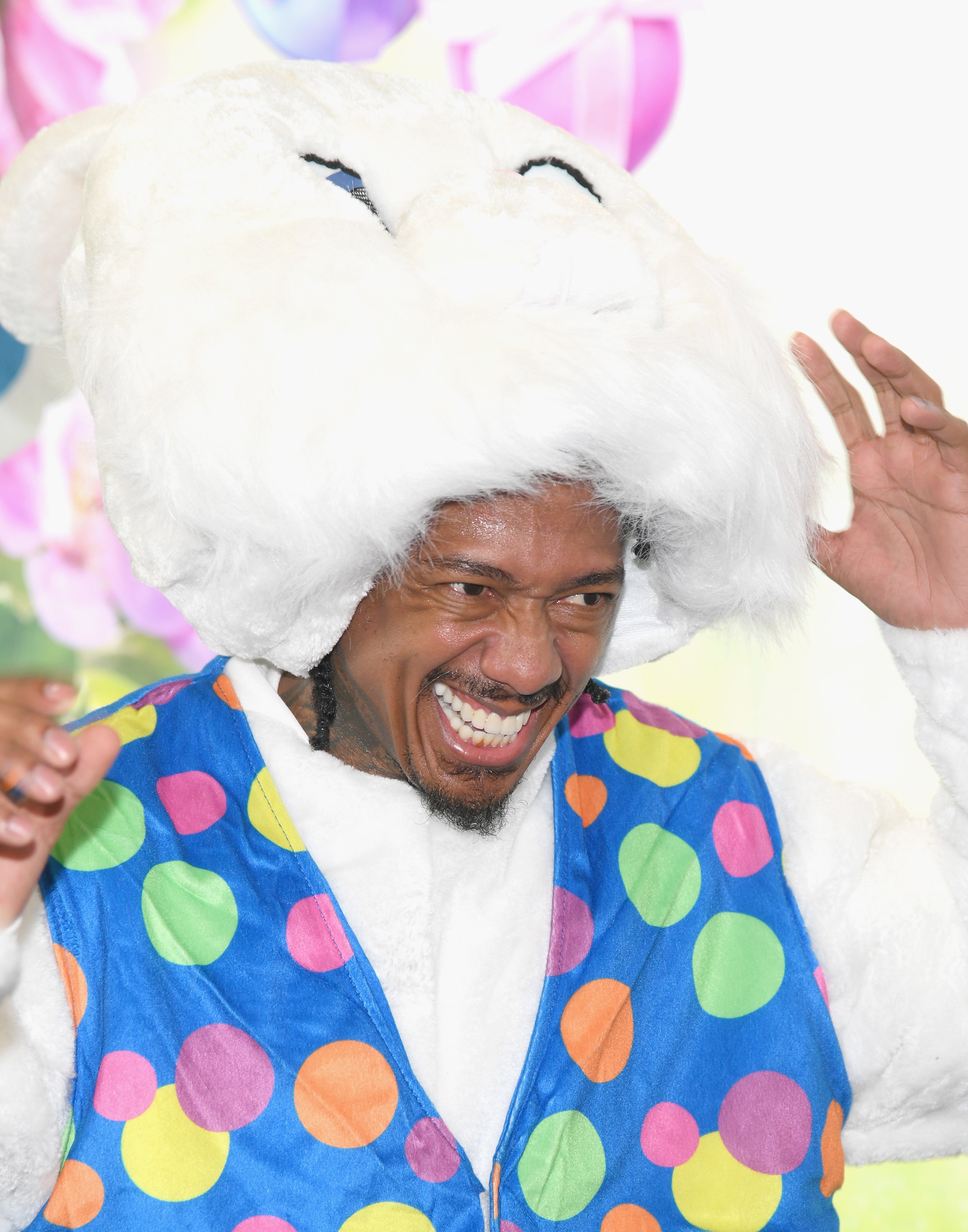 Nick Cannon dressed in a colorful polka dot vest and a large fluffy bunny hat, smiling and holding the hat