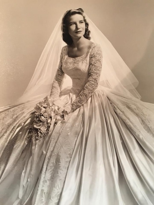 Woman in vintage wedding dress with lace sleeves and a long veil, holding a bouquet of flowers while sitting. Names unknown