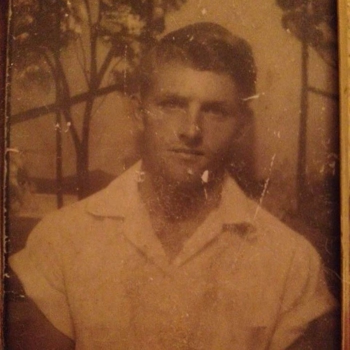 A vintage photograph of a man with short hair, wearing a button-up shirt, posing in front of a backdrop with trees