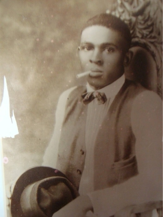 A historical portrait of a man with a short haircut wearing a bow tie, vest, and white shirt, holding a hat and a cigarette in his mouth. Names unknown