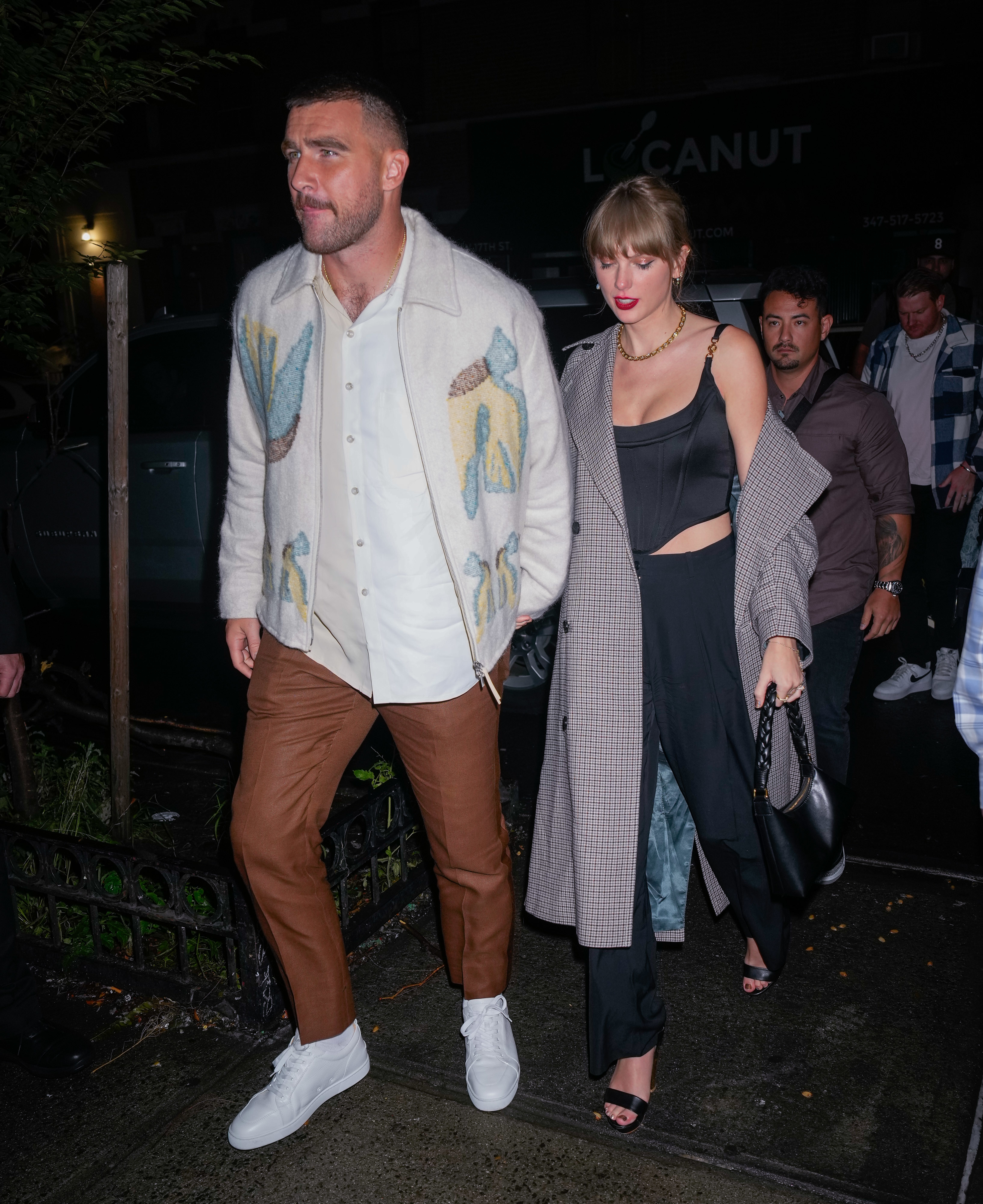 Taylor Swift in a stylish dark outfit and a coat walking with Travis Kelce, wearing a shirt, patterned jacket, pants, outside at night