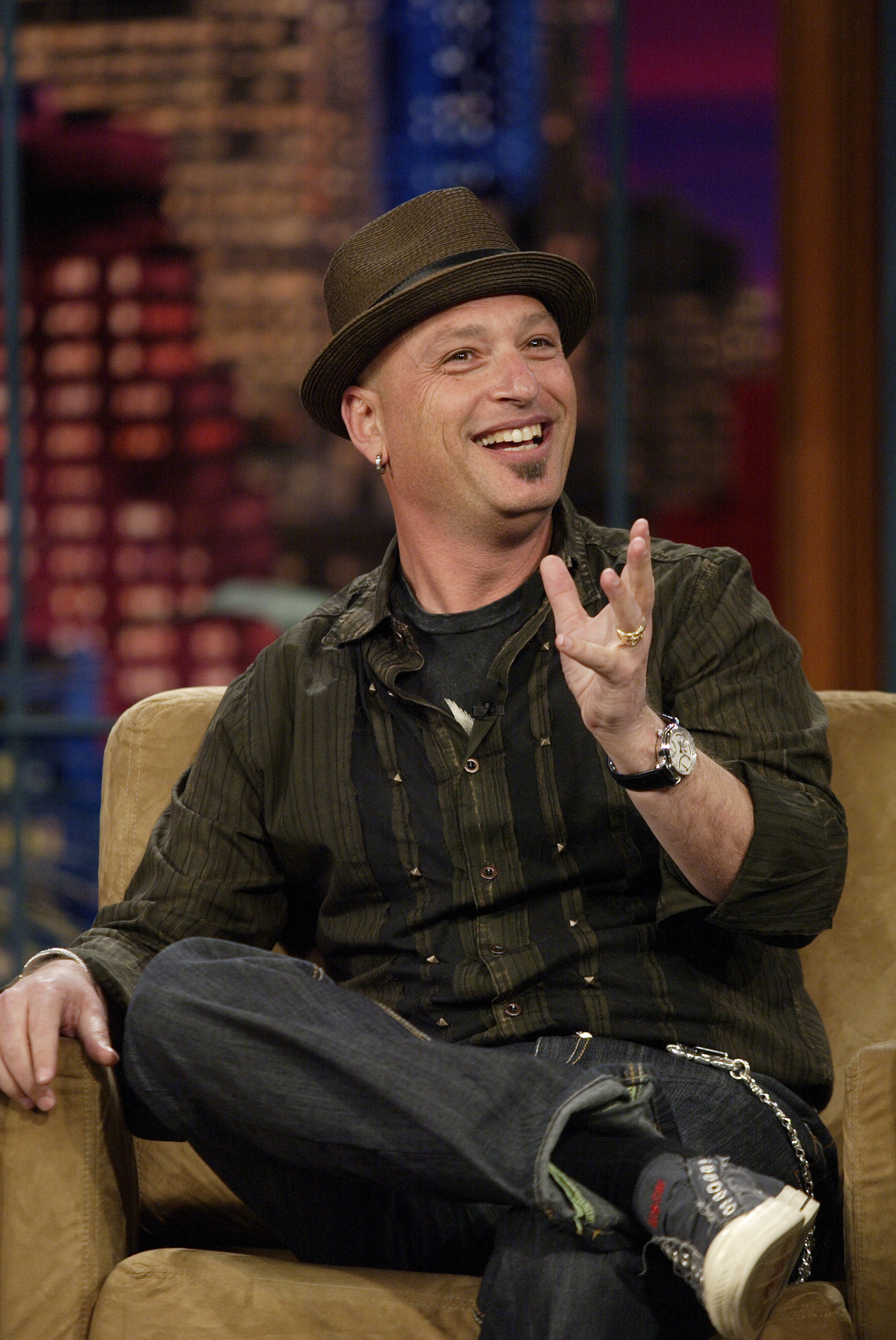 Howie Mandel, wearing a fedora and casual clothing, sits on a couch smiling.