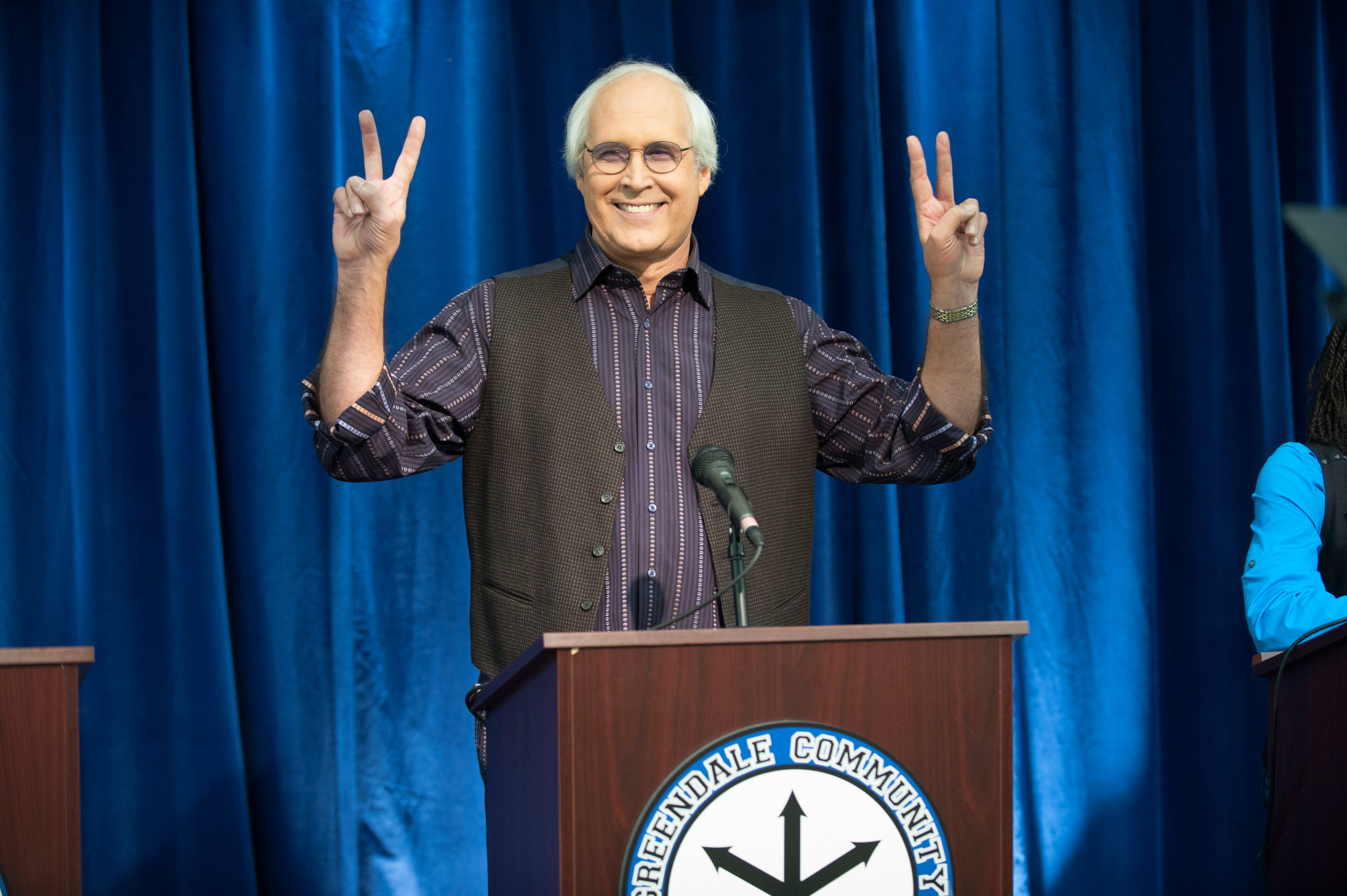 Chevy Chase on stage at a Greendale Community College event, smiling and holding up both hands with peace signs, wearing a button-up shirt and vest