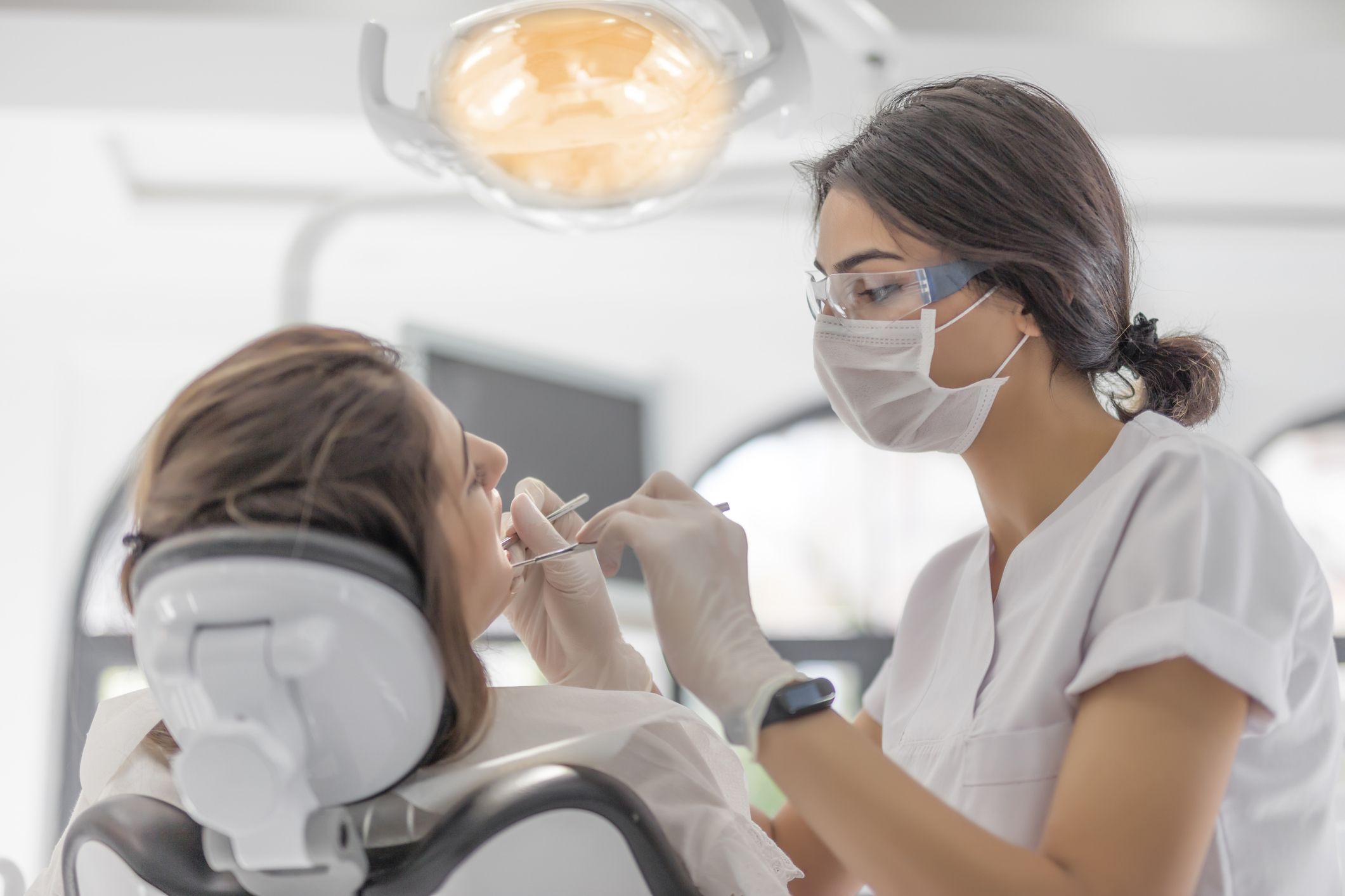 A dental hygienist is cleaning a patient&#x27;s teeth in a modern dental office. Both are wearing protective masks