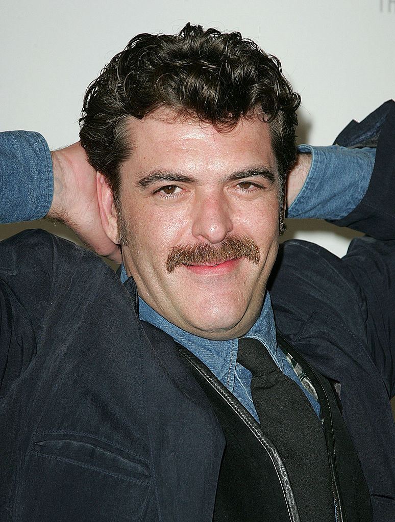 A man with a mustache smiles with his hands resting behind his head, wearing a denim shirt, dark jacket, and tie