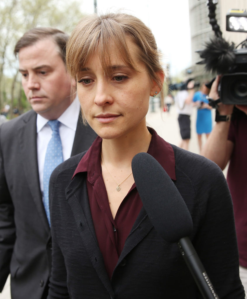 A woman, wearing a burgundy top and black cardigan, is facing the camera with a microphone in front of her. A man in a suit stands behind her