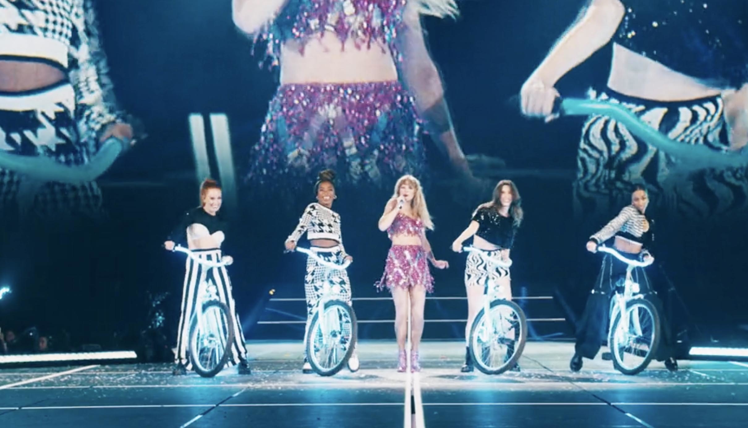 Taylor Swift performs on stage in a sparkling outfit, flanked by four dancers with illuminated bicycles during a concert