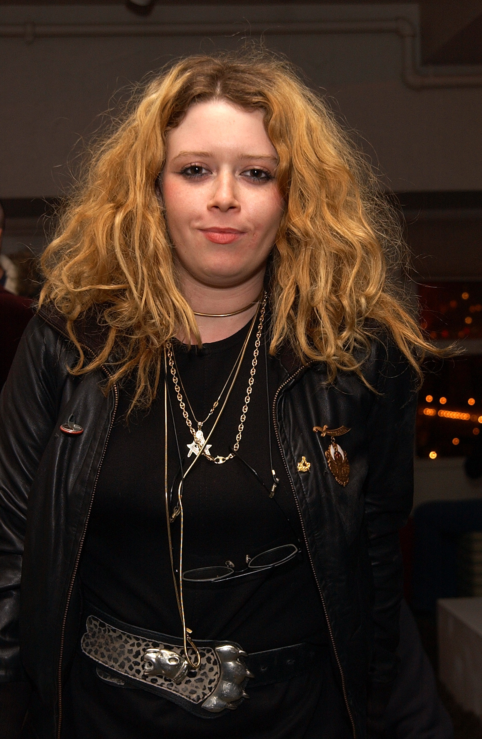 A young Natasha Lyonne is wearing a leather jacket, layered necklaces, and a wide belt