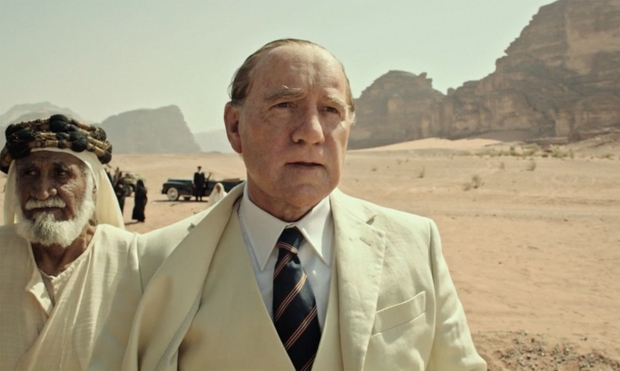 An elderly man in a beige suit and tie stands in a desert with rocky mountains in the background. Another man in traditional attire stands behind him