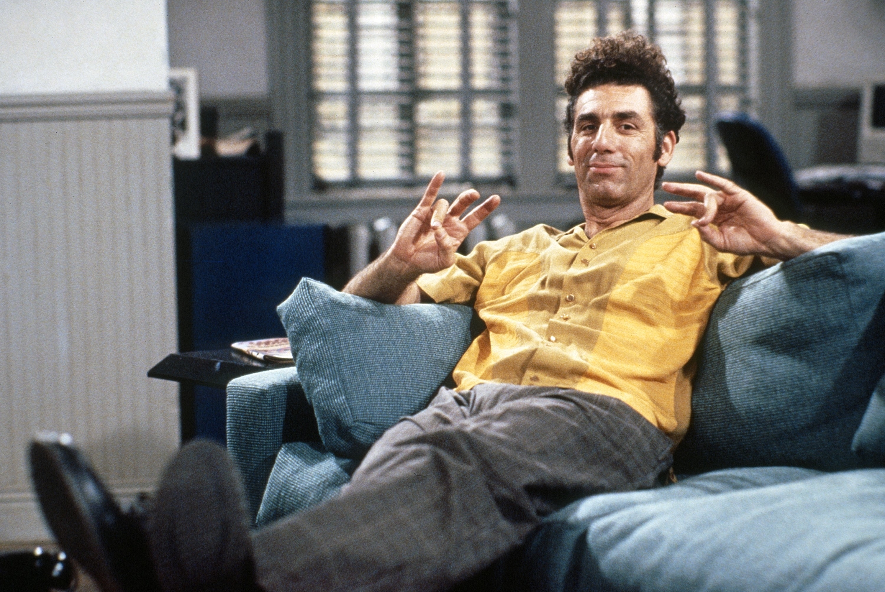 Michael Richards, as Kramer from Seinfeld, reclines on a couch with his legs up, wearing a casual shirt and relaxed trousers, holding a quirky hand gesture