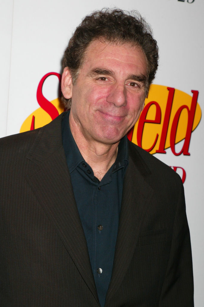 A person smiles at an event with a background featuring the &quot;Seinfeld&quot; logo. They are wearing a dark suit and a navy shirt