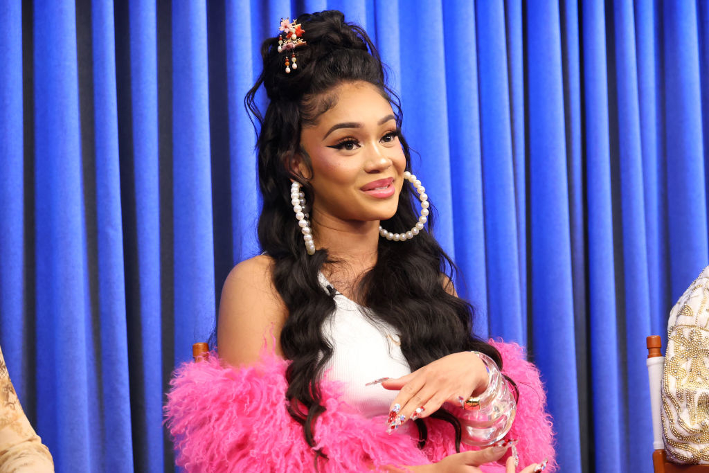 Saweetie sits in front of a blue backdrop, wearing a white top with a pink fluffy coat, large pearl hoop earrings, and her hair styled in long waves