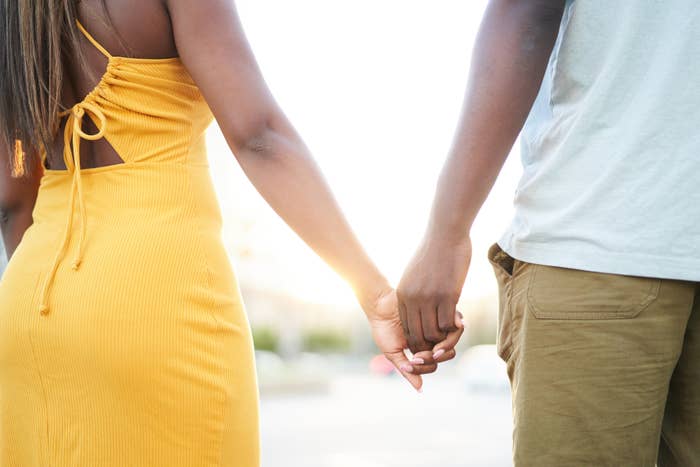 A woman in a sleeveless dress and a man in casual clothes are shown from behind, holding hands while walking in a bright outdoor setting