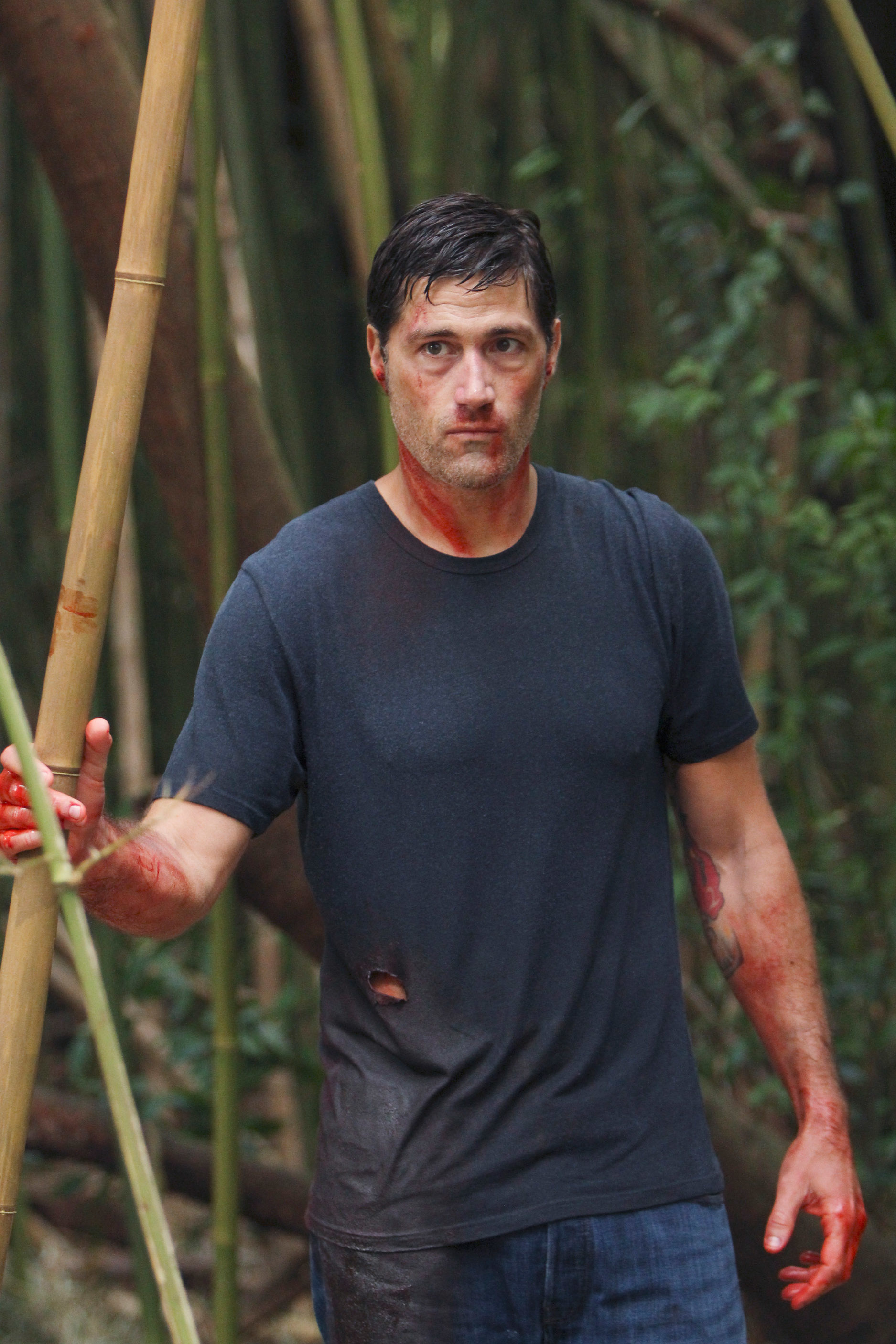 Matthew Fox in a distressed t-shirt, gripping bamboo stalks in a forest setting, with blood on his hands and shirt